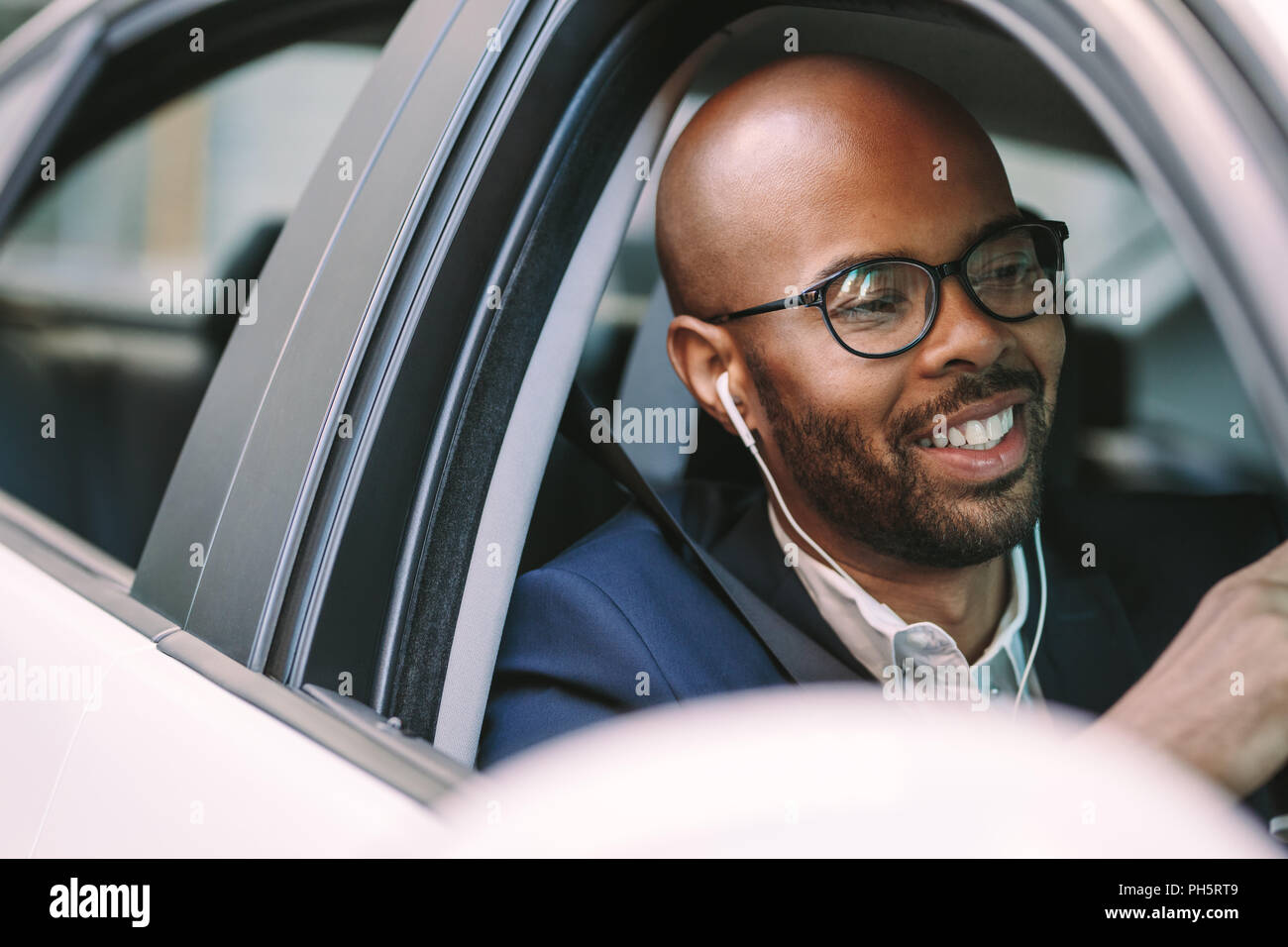 Giovane uomo calvo sorridente dentro la macchina durante la guida. L'uomo africano in tuta e indossare gli auricolari godendo di guida di un'auto. Foto Stock