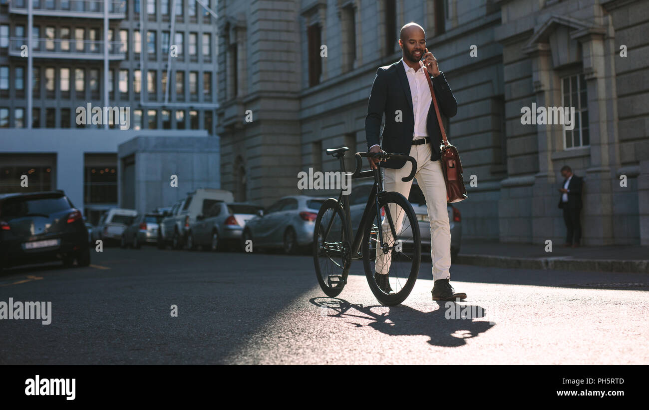 Giovane imprenditore passeggiate all'aperto con una bicicletta e utilizzando il telefono cellulare. L'uomo africano di andare al lavoro in bicicletta a parlare su telefono cellulare sulla strada della citta'. Foto Stock