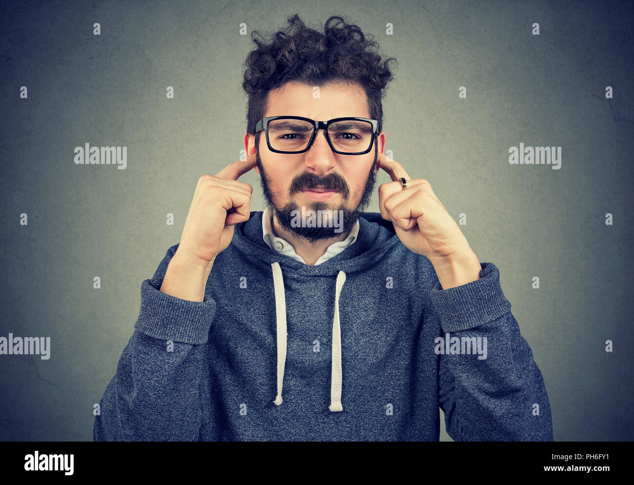 Infastiditi giovani hipster man looking grumpy in telecamera e coprendo le orecchie dal rumore evitando rumori forti e situazione di conflitto su sfondo grigio Foto Stock