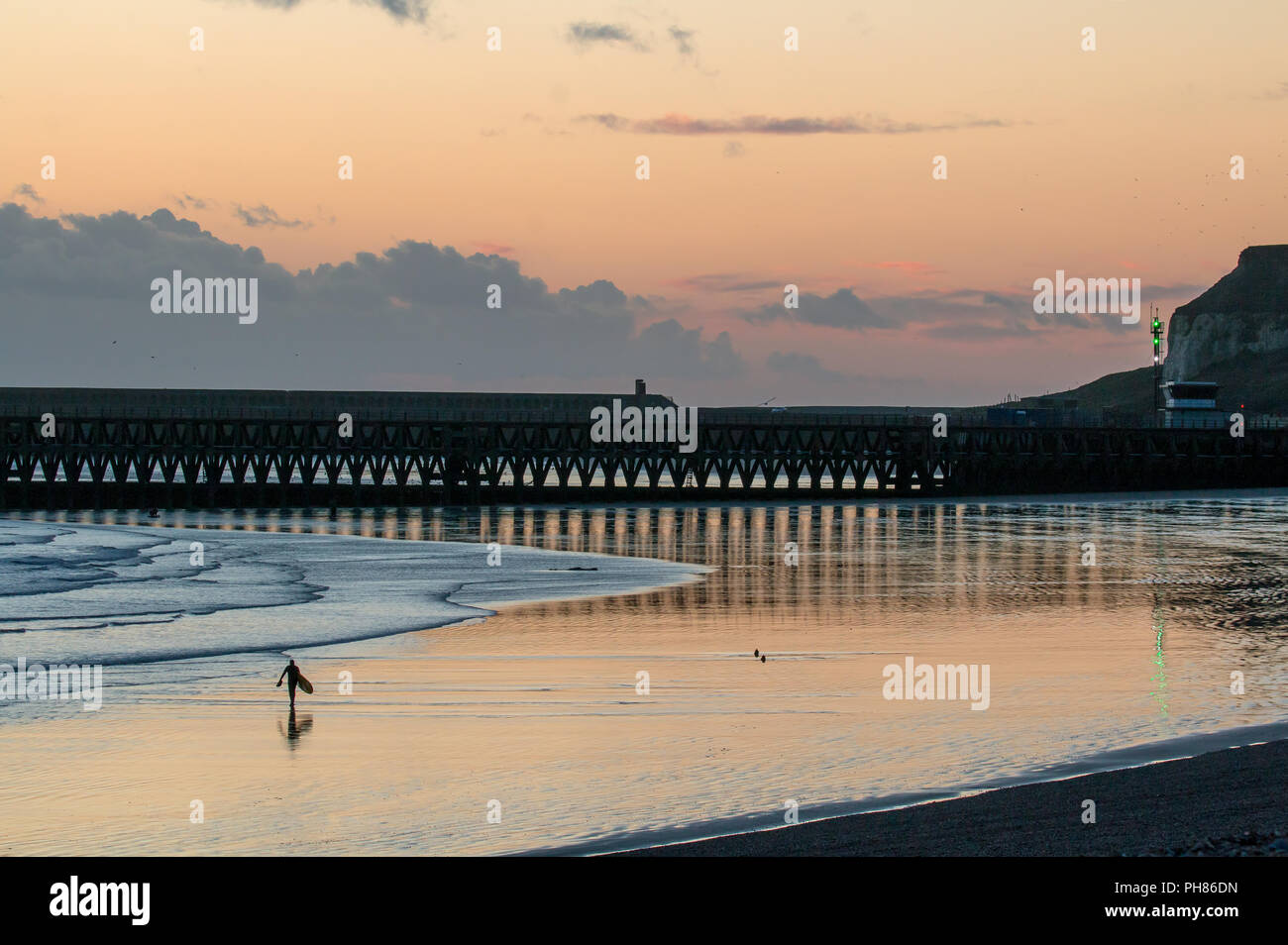 Persona non identificabili con la tavola da surf a piedi a bassa marea su Seaford spiaggia al tramonto, con riflessioni di Newhaven Harbour sulla sabbia bagnata. Foto Stock