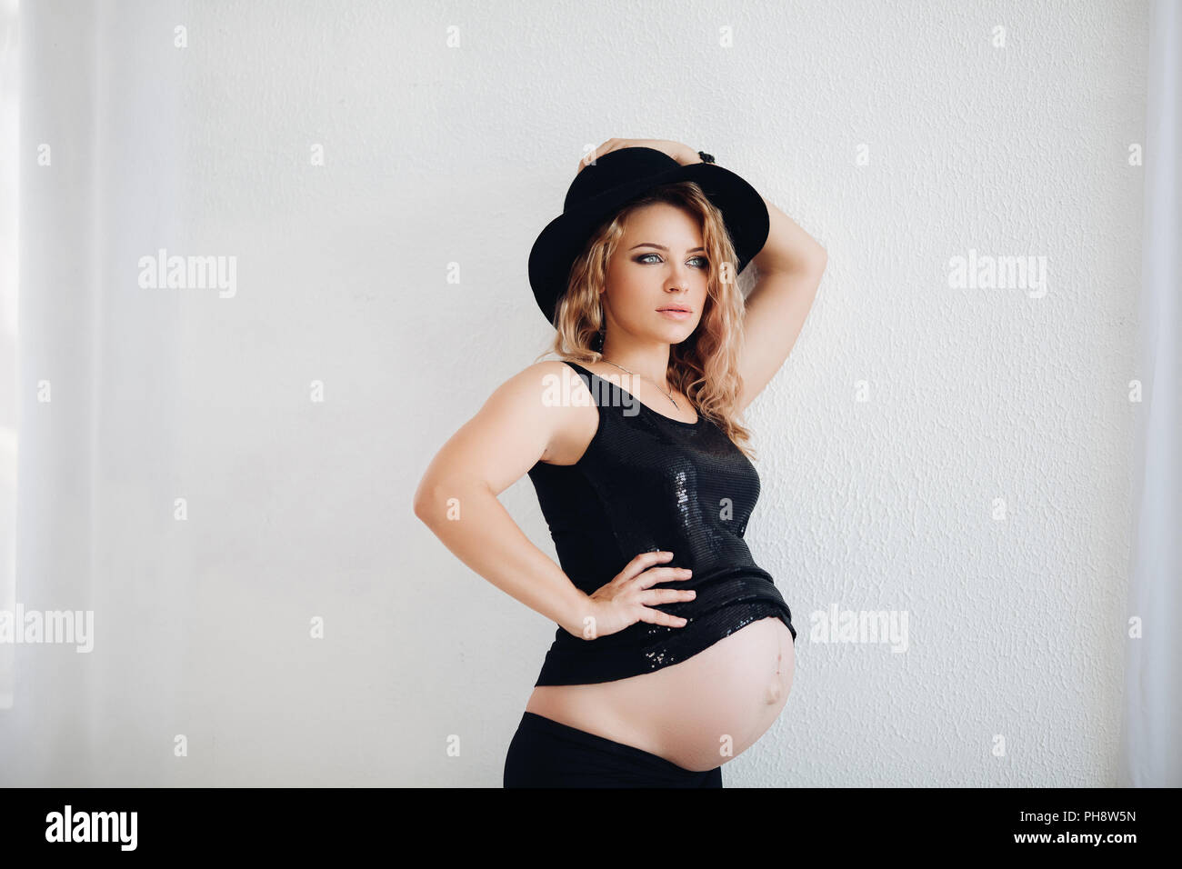 Elegante donna incinta in black hat e frizzante top abbracciando il suo ventre. Foto Stock