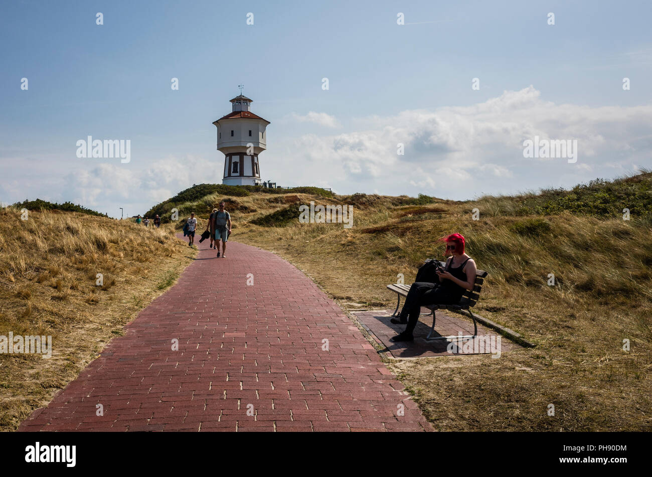 Water Tower, Langeoog. Germania, Deutschland. Un sentiero conduce verso una attrazione turistica, l'iconica white water tower - Wasserturm. Una giovane donna con i capelli rossi, si siede su un banco di pubblico per controllare il suo telefono cellulare. L'erba sulle dune di sabbia sono stati sbiancati dal sole durante l'estate di onda di calore. Foto Stock