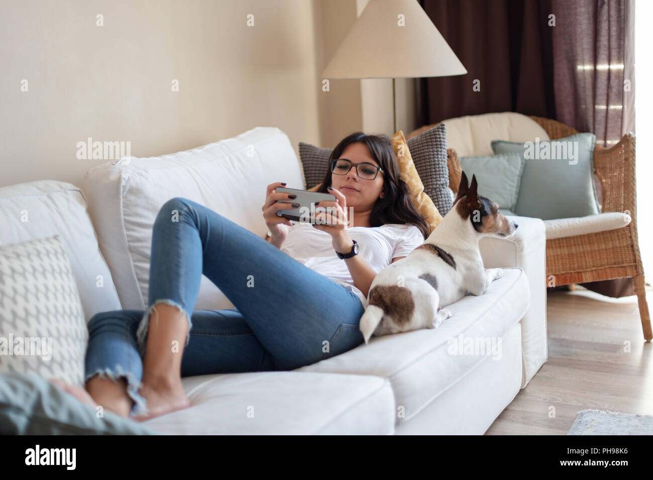 Attraente giovane donna la condivisione di un divano con il suo cagnolino che giace accanto a lei come usa il suo telefono cellulare in luoghi chiusi a casa Foto Stock