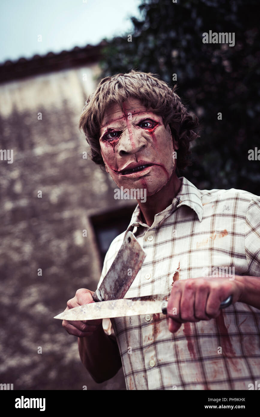 Un pauroso sfigurato uomo affilatura di un arrugginito e sanguinosa cleaver con un arrugginito e coltello insanguinato, di fronte a una casa in rovina in un inquietante paesaggio Foto Stock