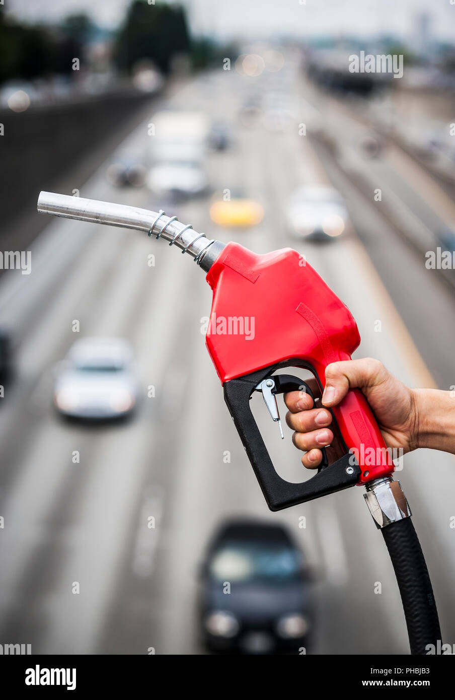 Una mano che tiene un gas ugello della pompa al di sopra di un fuori fuoco autostrada scena. Foto Stock