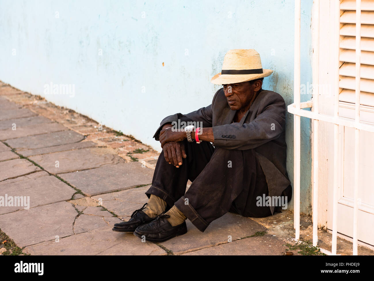 Trinidad / Cuba - Marzo 15, 2016: indigenti uomo cubano, indossando la paglia fedora e ben indossato tuta, siede sulla terra contro la parete blu in plaza square. Foto Stock