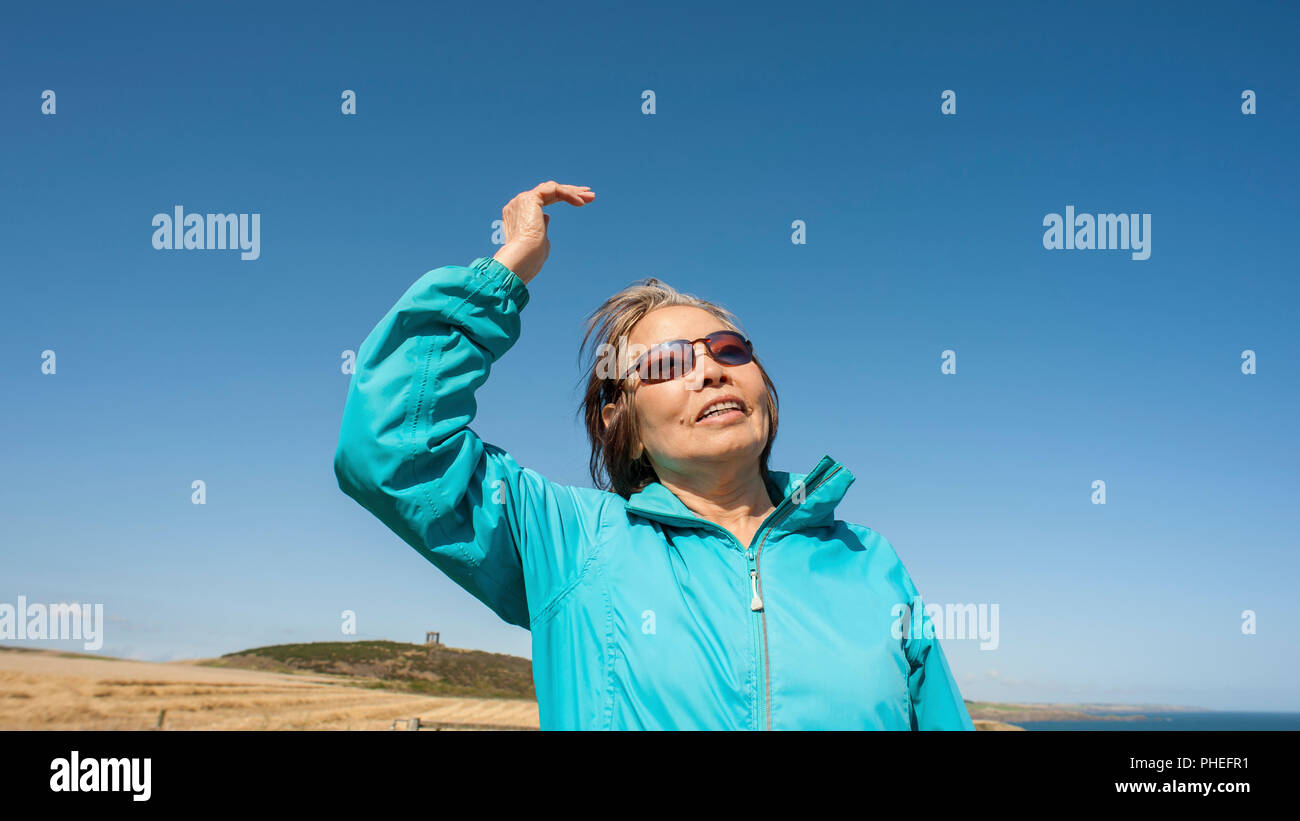 Matura, Asian lady solleva la mano destra come si guarda lontano nel cielo blu con espressione facciale per essere interpretato. Foto Stock