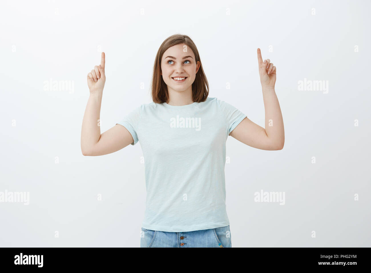 Mi chiedo che cosa nascondere verso l'alto. Intrigato e gioiosa upbeat femmina con un ampio sorriso curioso di puntamento e di sguardo fino in piedi in t-shirt casual e jeans oltre il muro bianco promozione di raffreddare lo spazio di copia Foto Stock