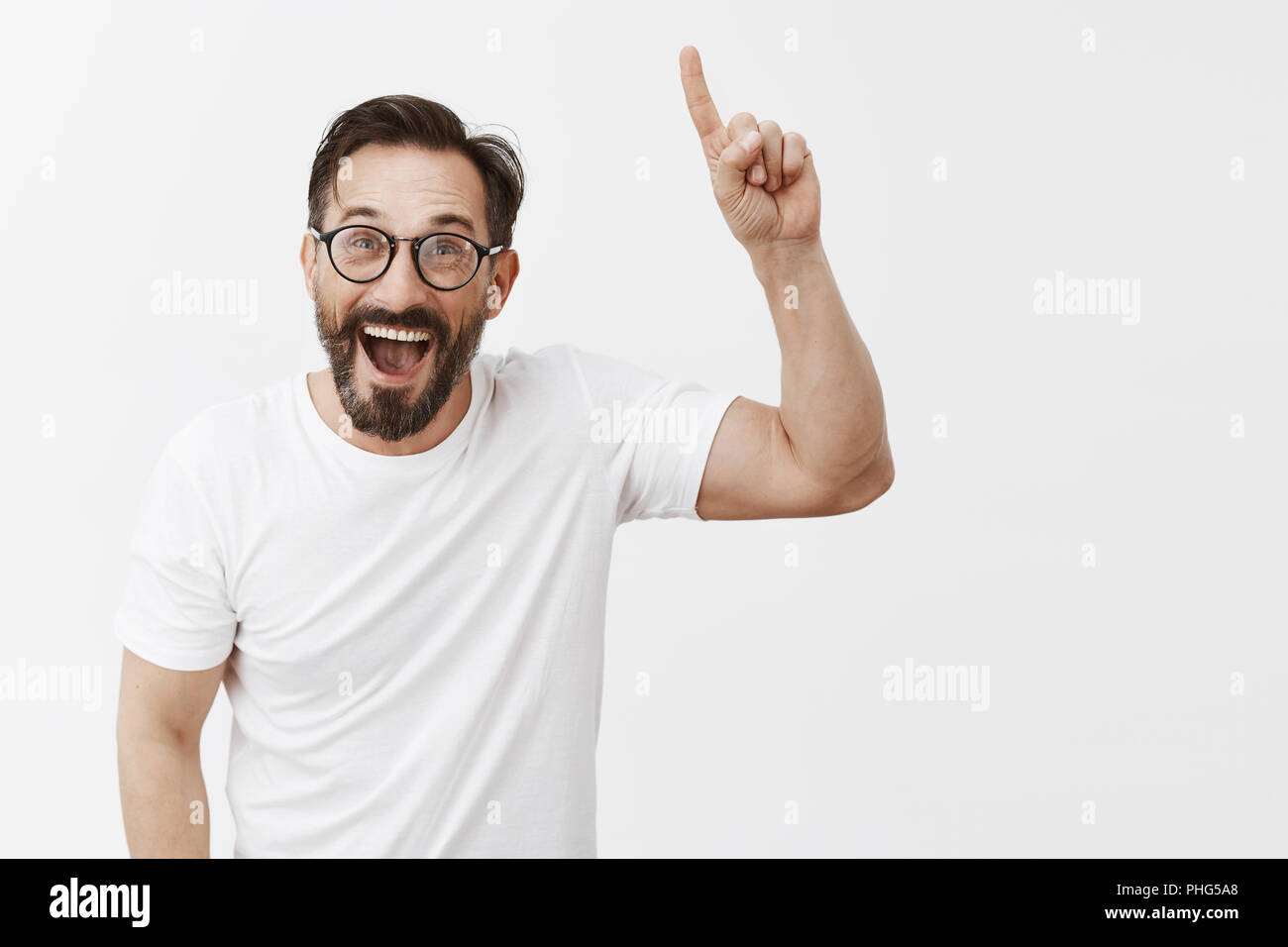 Emozionato felice uomo bello con la barba in occhiali trendy e t-shirt, sollevando il dito indice per puntare in alto nel cielo o mostra il gesto di Eureka, sorridenti pieni di gioia e di allegrezza e trionfante, essendo a destra Foto Stock