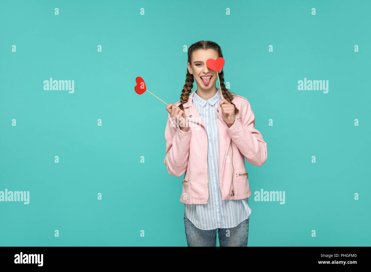 Funny bella ragazza in stile casual, pigtail pettinatura e camicia rosa, permanente e la holding cuore rosso adesivi e guardando la fotocamera con la lingua di fuori Foto Stock