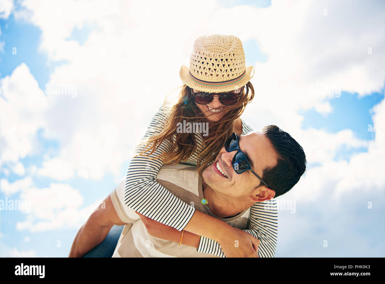 Bel giovane uomo con la sua amorevole moglie o fidanzata avendo divertimento rilassante per la vacanza estiva piggy back riding contro un soleggiato nuvoloso cielo blu Foto Stock