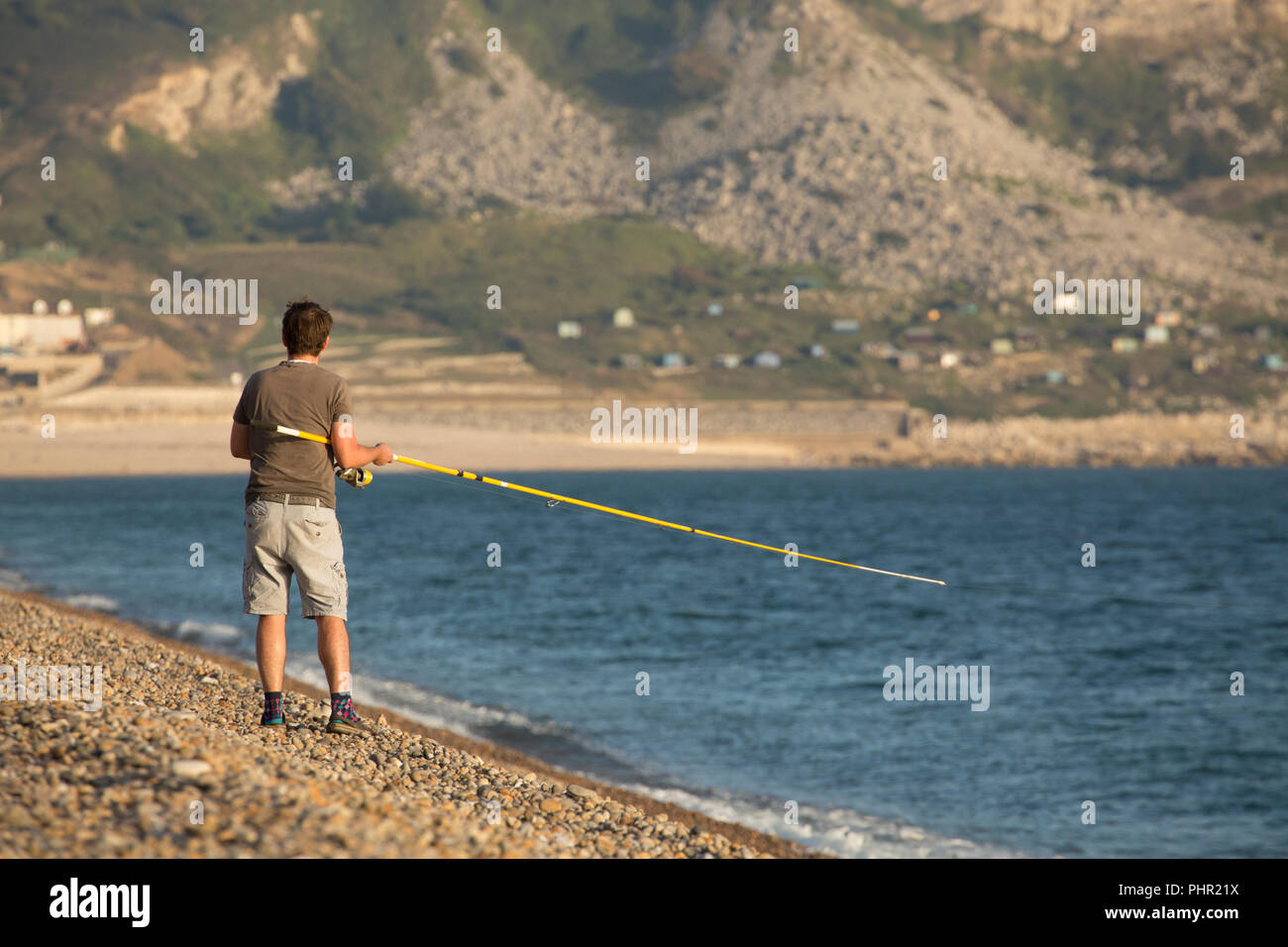 Un pescatore che pesca nel tardo pomeriggio per sgombro su Chesil Beach contro uno sfondo di Chesil Cove e le scogliere dell'isola di Portland. Il Dorset Foto Stock
