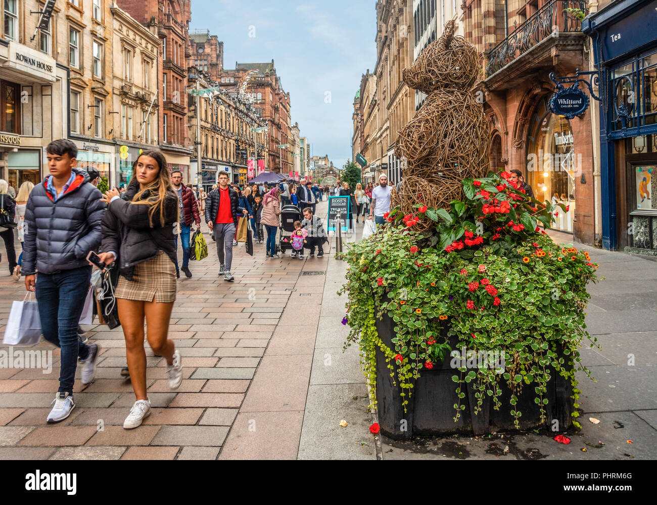 Gli amanti dello shopping nella zona pedonale di Buchanan Street nel centro di Glasgow, Scozia. Un vimini orsacchiotto scultura su una piantatrice di fiore in primo piano. Foto Stock