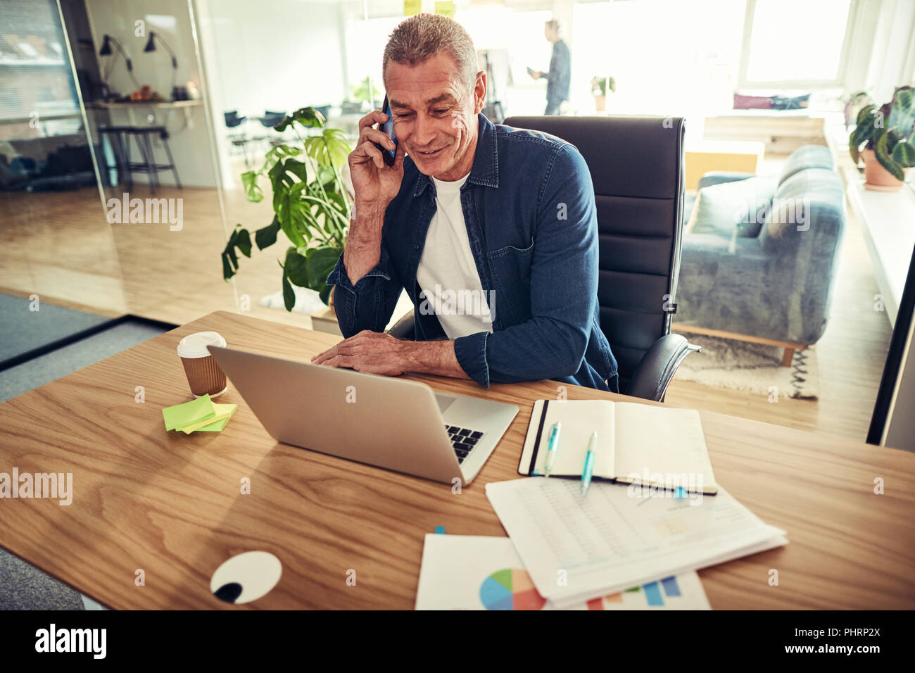 Sorridente imprenditore maturo parlando sul suo cellulare e lavorare online con un computer portatile mentre è seduto alla sua scrivania in un ufficio Foto Stock