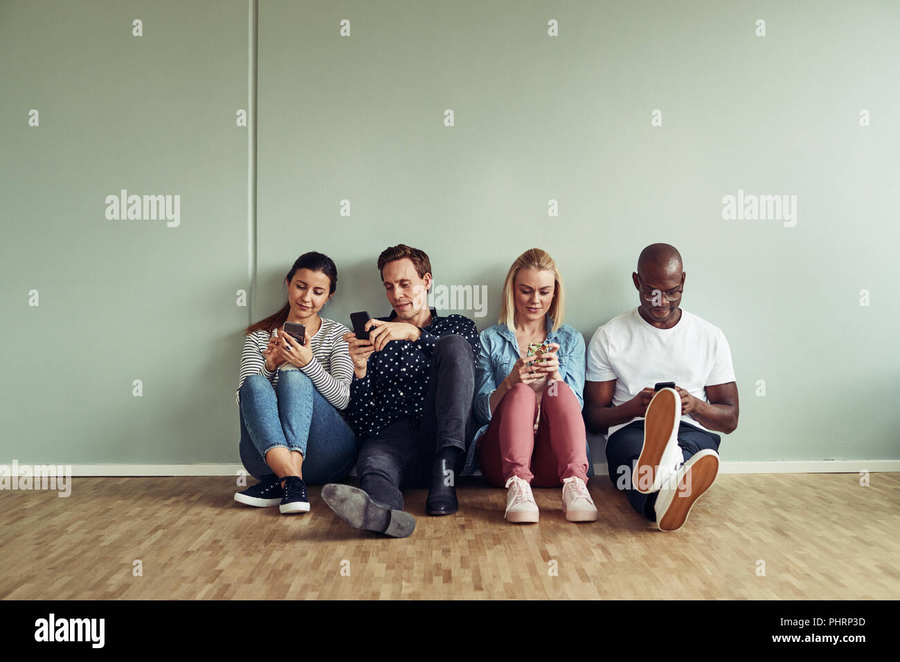 Gruppo di diversi giovani colleghi di lavoro seduti insieme su un pavimento di Office utilizzando i loro telefoni cellulari durante l'aperitivo Foto Stock