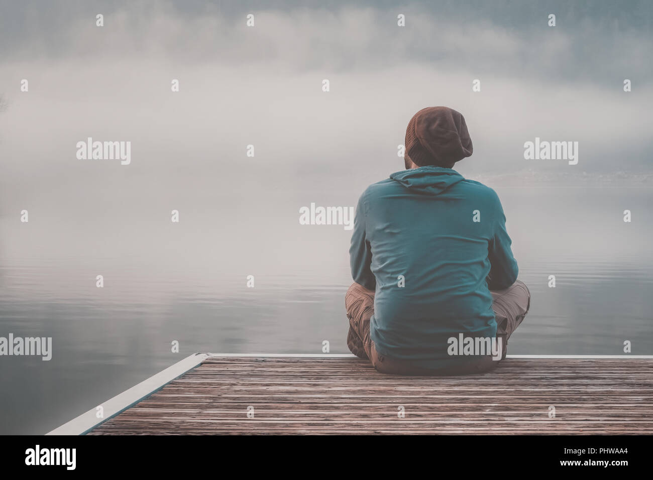 Giovane uomo con cappa di seduta sul molo di un lago in un giorno di nebbia. Foto Stock