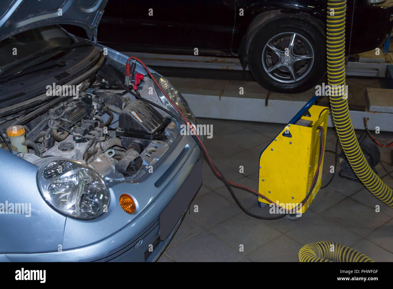 Il giallo caricabatterie carica della batteria auto di colore blu con un cofano aperto nell'officina di riparazione con un SUV nero in piedi su un ascensore per la riparazione Foto Stock