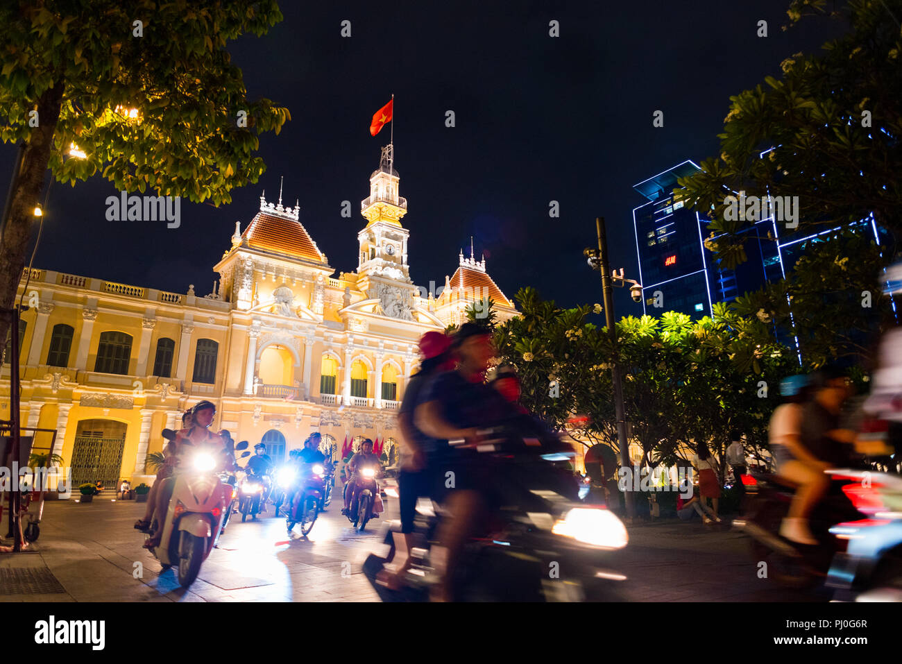 La città di Ho Chi Minh, Vietnam - Maggio 1, 2018: la gente guida scooter con i fari accesi passando da Saigon City Hall, Centro Vincom torri e alberi di notte Foto Stock