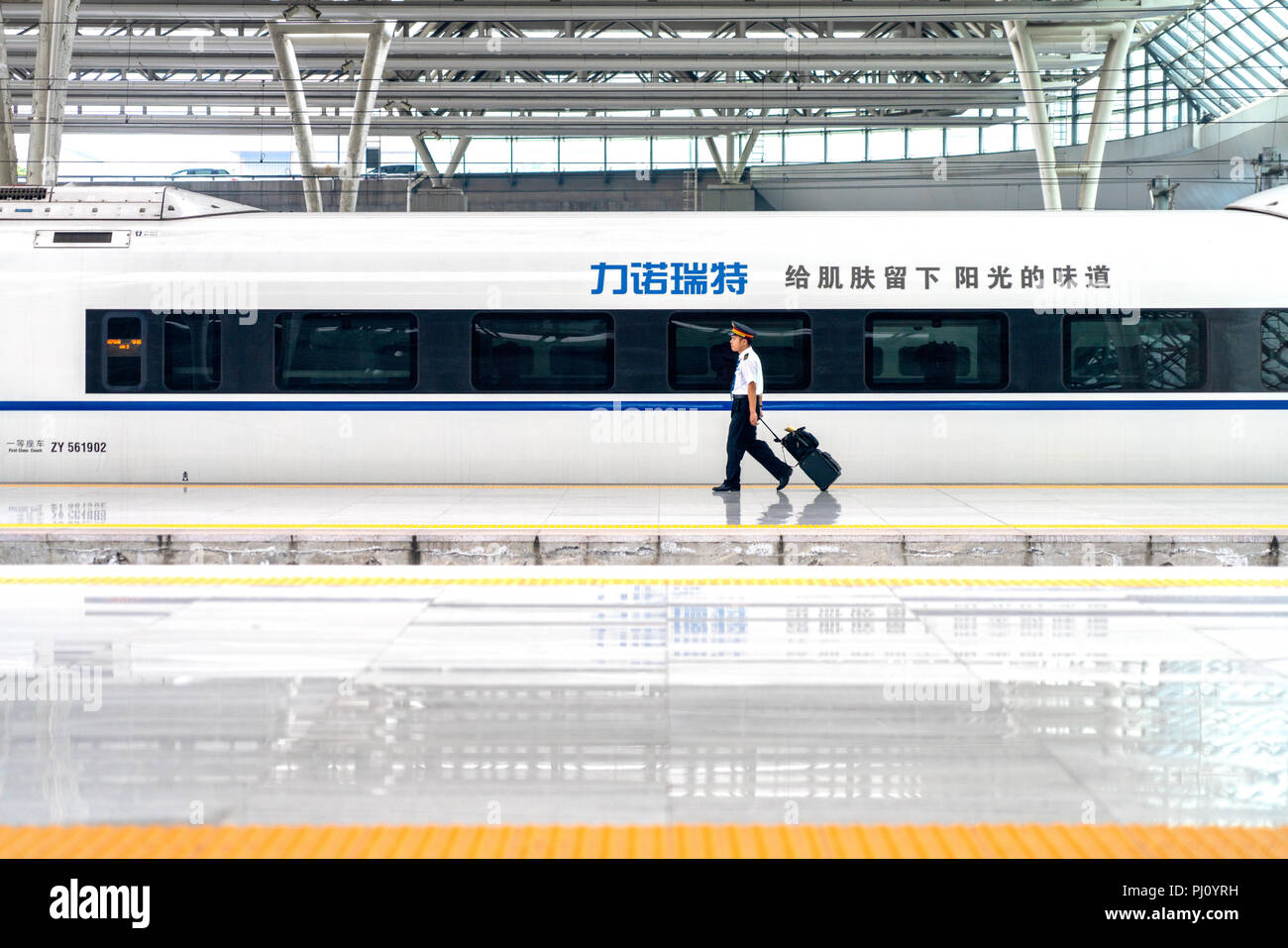Un asiatico il treno ad alta velocità conduttore passeggiate lungo la piattaforma con un treno auto dietro di lui, mentre si tira la sua valigia in Cina a Shanghai. Foto Stock