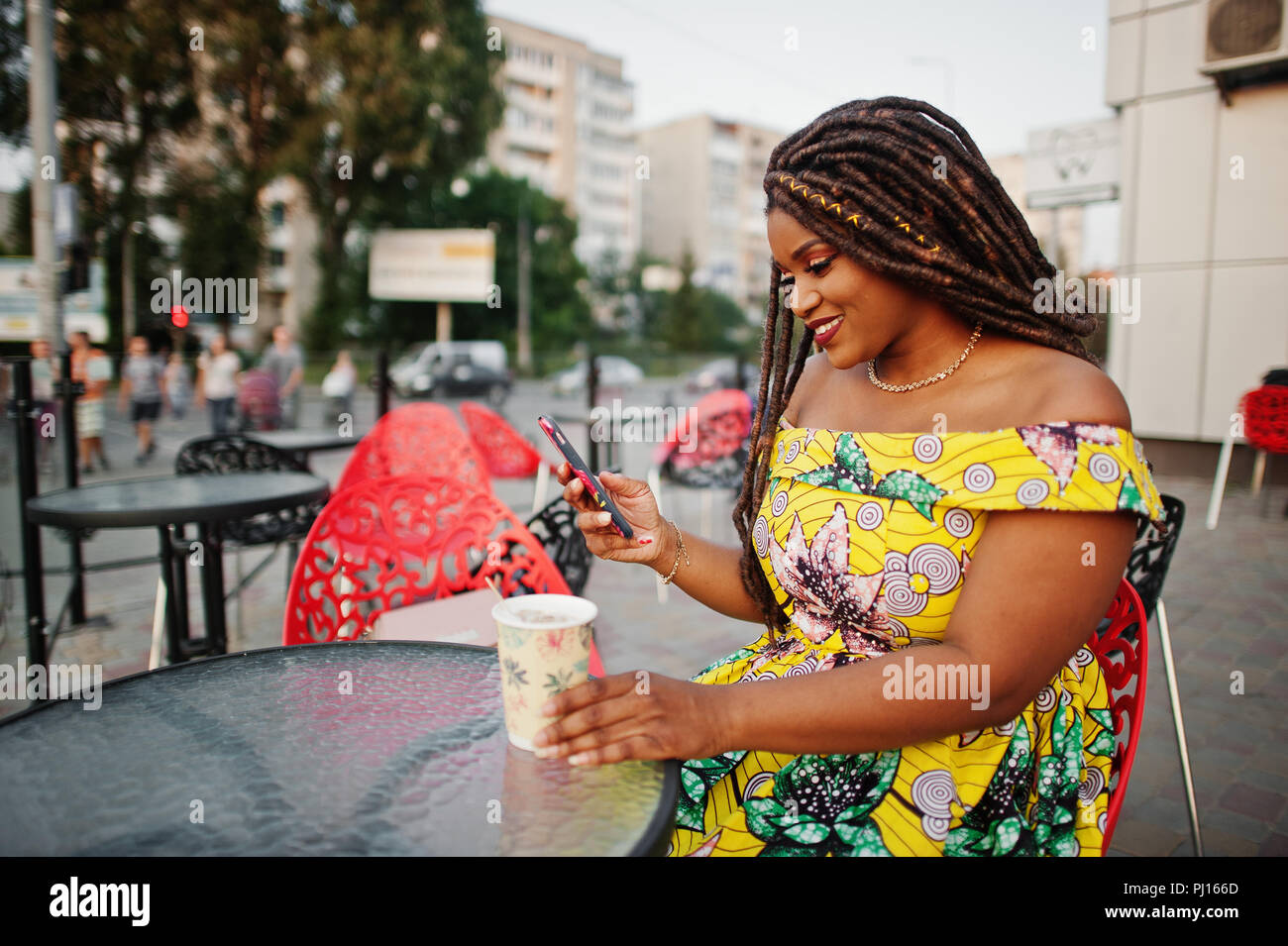 Carino piccola altezza African American Girl con Dreadlocks, usura in corrispondenza di colore giallo abito, seduti al cafe esterno sulla sedia rossa e di bere il caffè. Foto Stock