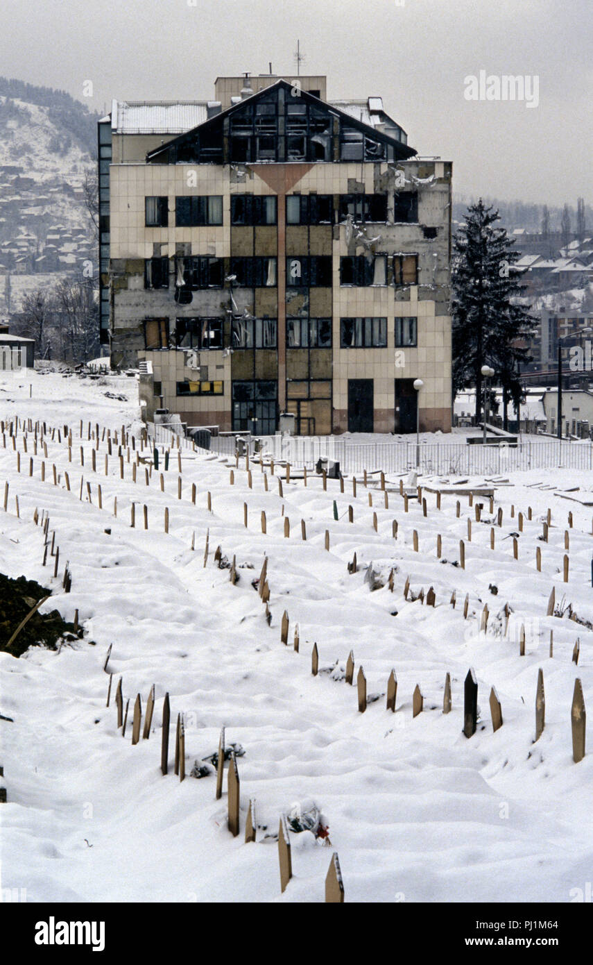 4 Marzo 1993 durante l'assedio di Sarajevo: un gravemente danneggiato edificio sorge al di sotto di ranghi di coperte di neve bosniaci musulmani tombe nel cimitero di Lion, appena al di sotto dell'Ospedale Kosevo. Le tombe sono tutti datati 1992. Foto Stock