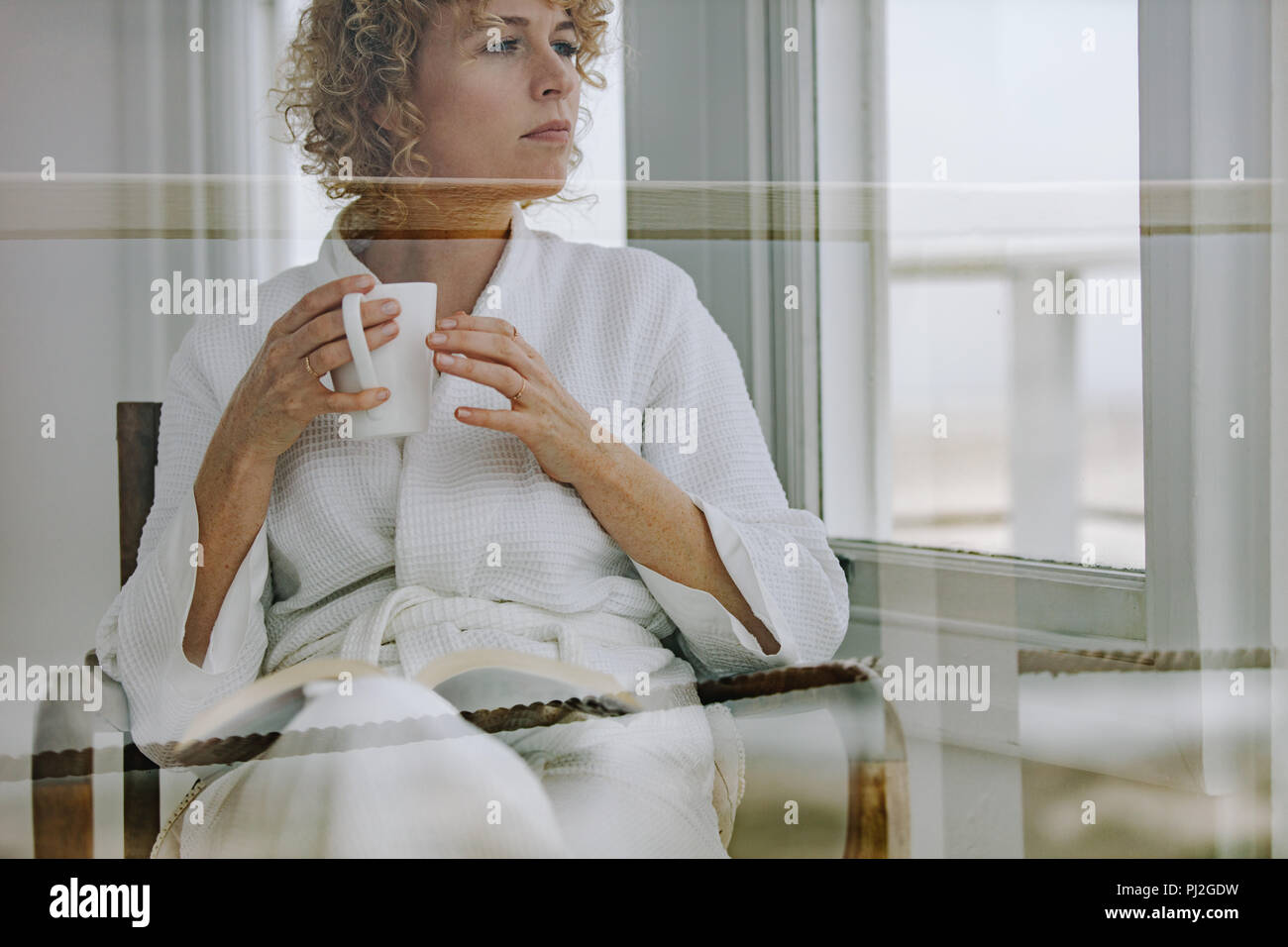 Donna in accappatoio leggendo un libro gustando un caffè. Donna che guarda fuori dalla finestra e pensare durante la lettura di un libro. Foto Stock