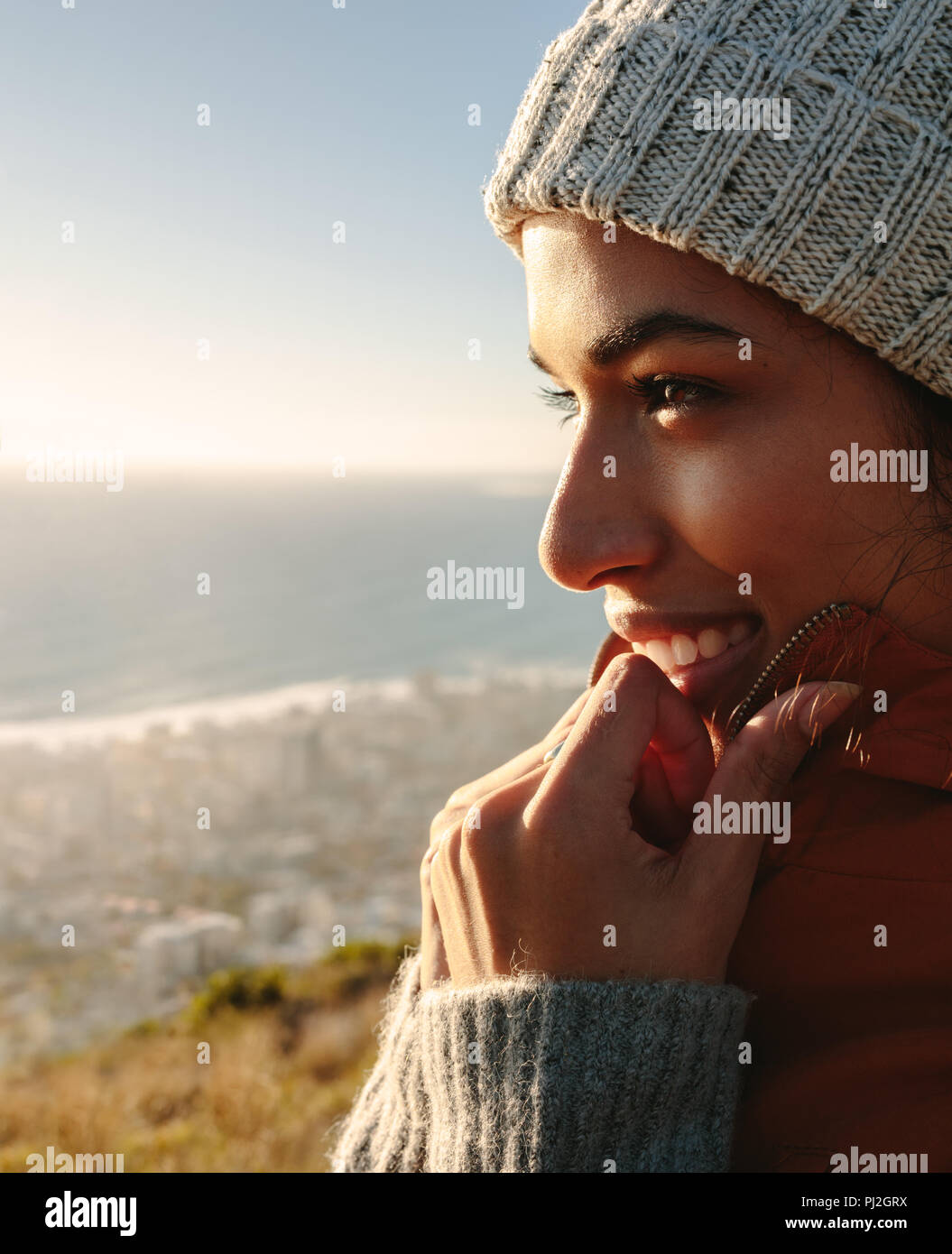 Close up di sorridere donna africana godendo della splendida vista del mare dalla cima della montagna. Femmina in una giacca calda e cappuccio a maglia. Foto Stock