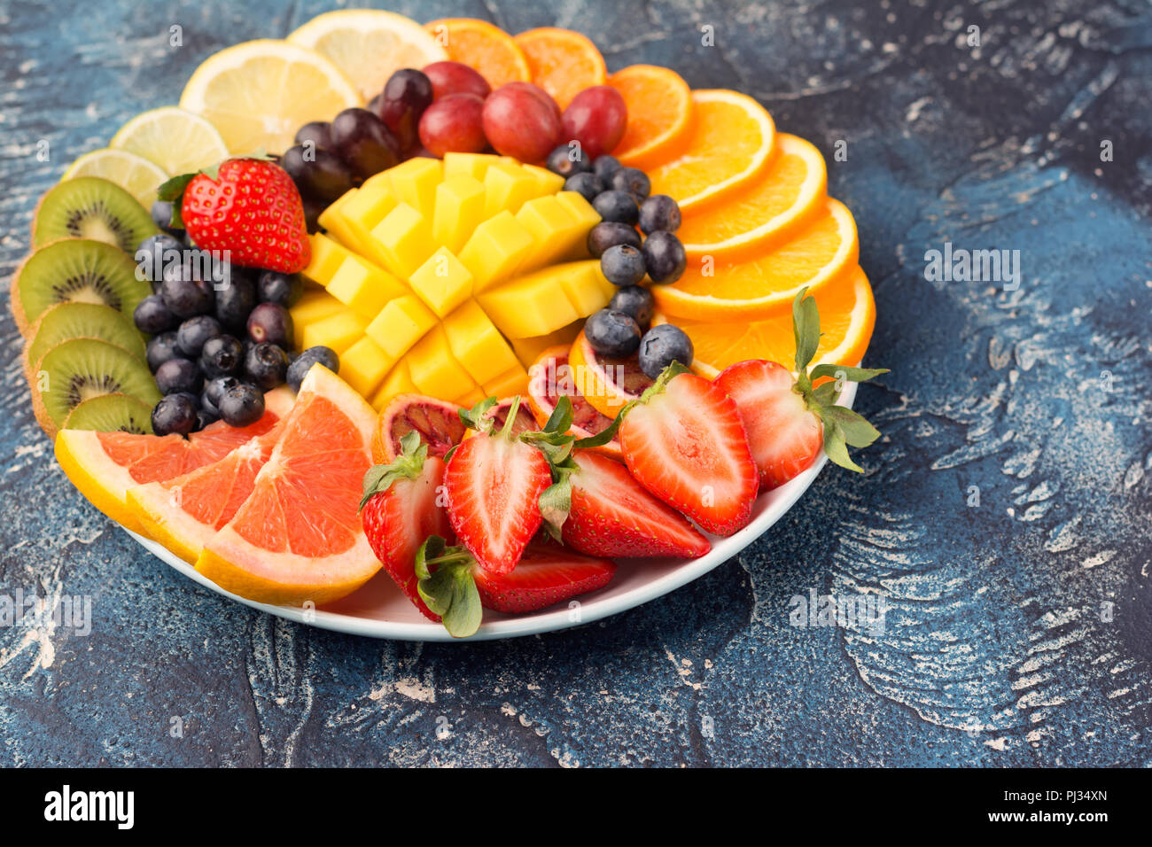 Sani frutti e bacche platter nei colori dell'arcobaleno close up, fragole, mango, uva, arance e kiwi, limoni sul tavolo blu, copiare lo spazio per testo, il fuoco selettivo sul mango Foto Stock
