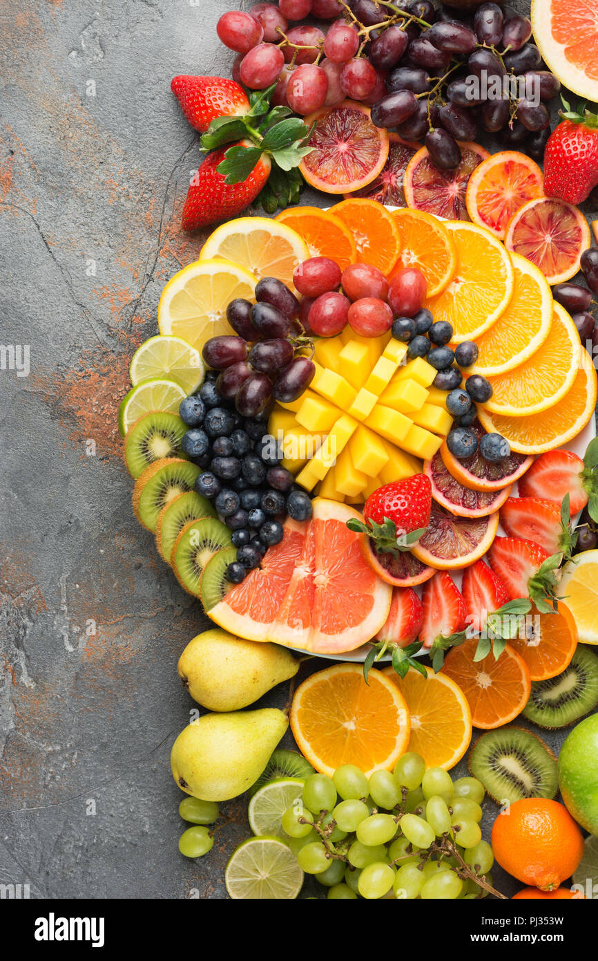 Assortimento di frutta tagliata nei colori dell'arcobaleno Arance Uve mango fragole kiwi mirtilli pompelmo sul cemento grigio tabella, vista dall'alto, copia di spazio, il fuoco selettivo Foto Stock