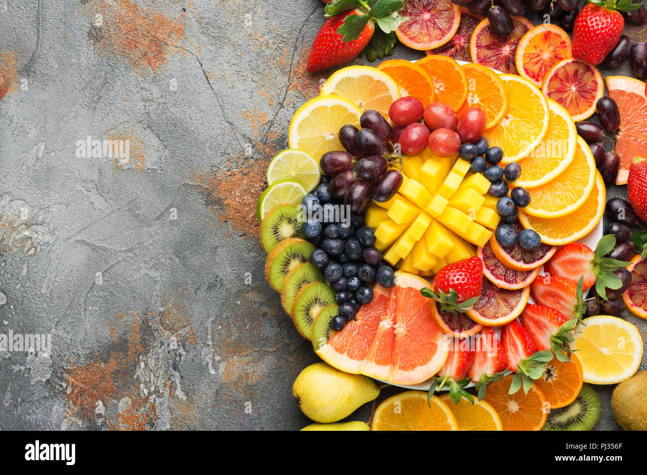 Colorata frutta tagliata nei colori dell'arcobaleno Arance Uve mango fragole kiwi mirtilli pompelmo sul cemento grigio tabella, vista dall'alto, copia di spazio, il fuoco selettivo Foto Stock