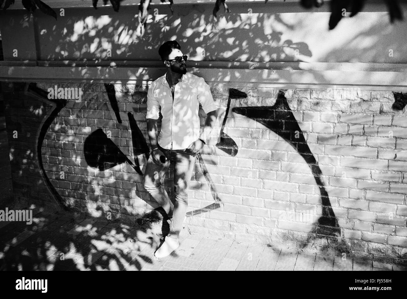 Elegante tall arabian modello da uomo in camicia bianca e jeans e occhiali da sole poste lungo la strada della citta'. Barba attraente ragazzo arabo contro il muro di graffiti. Foto Stock