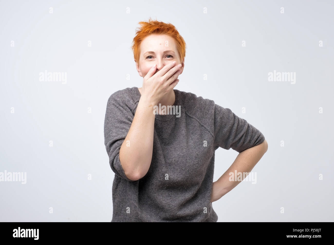 Funny donna matura con corti capelli rossi chiude la sua bocca con palm audizione divertente scherzo. Foto Stock