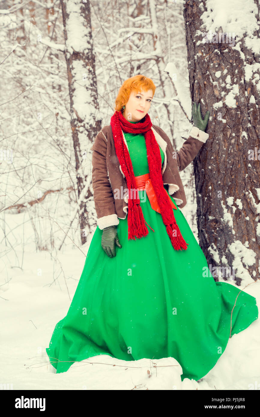 Fiery i capelli rossi donna in una palla abito verde con un rosso cintura di pelle in costume del nano assistant Santa Claus in inverno foresta con enormi cand Foto Stock