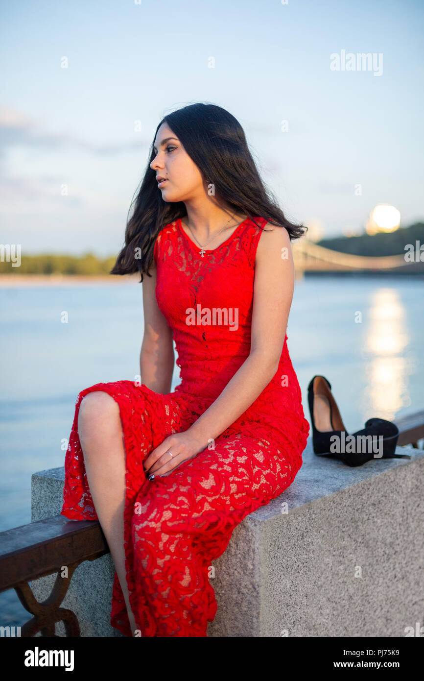 Una ragazza in un lungo abito rosso è seduto sulla recinzione di il greto del fiume. Foto Stock