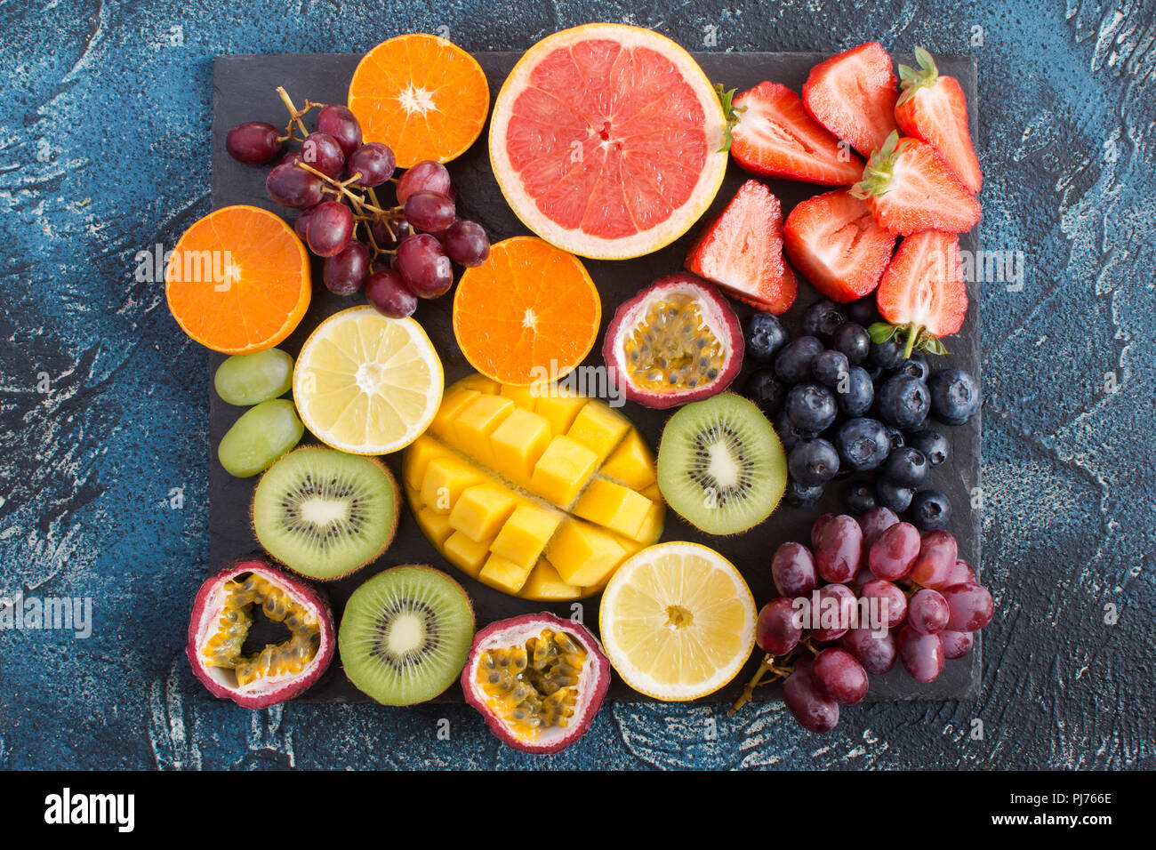 Materie frutta piatto: arance, limoni, kiwi, pompelmi, fragole, uva, mirtilli, frutto della passione, mango sul nero ardesia sul blu scuro b Foto Stock