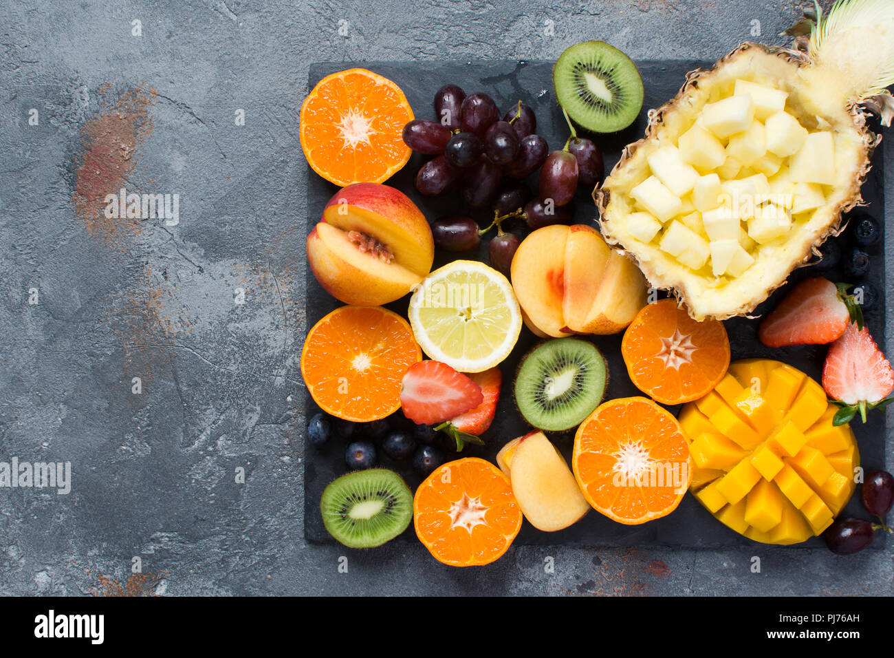 Materie frutti e bacche di piatto sulla pietra grigia sfondo, copia dello spazio. Ananas, mango, satsuma, kiwi, pesche, arance, fragole, uva, blu Foto Stock