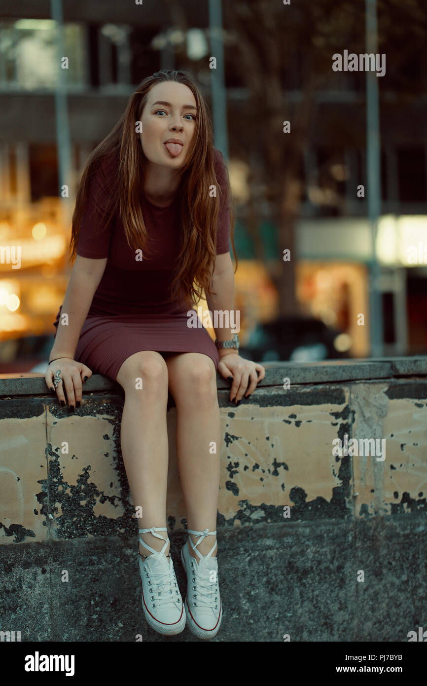 Colpo all'aperto di giovani piuttosto rossastra sorridenti ragazza che mostra la linguetta e divertirsi seduti sulla strada, indossare un abito bianco e stivali. Foto Stock