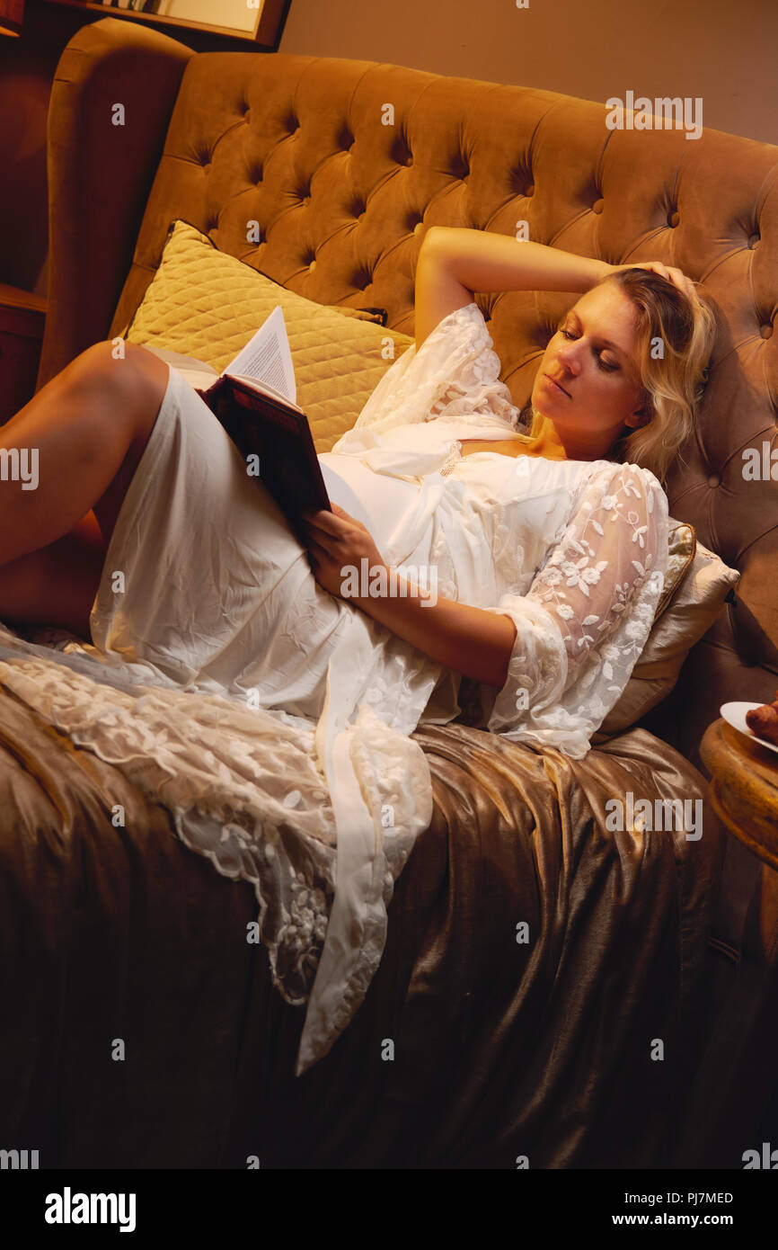 Incinta giovane donna bionda in abito di riposo sdraiato sul letto in camera da letto e la lettura di un libro sulla maternità o chicklit, donna romanzo a casa. Relax, Foto Stock