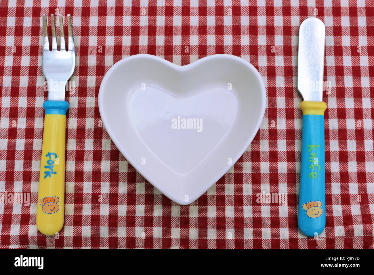 Un cuore a forma di ciotola di cibo e figli di coltello e forchetta su un rosso Gingham tovaglia. Tabella o impostazione di picnic. I bambini del posto dell'impostazione. Foto Stock
