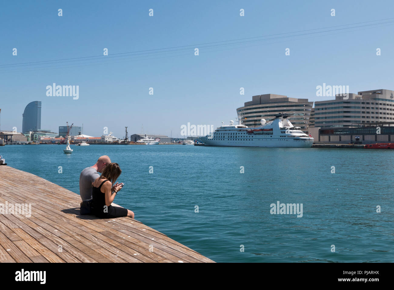 Un paio di posa su pavimento di legno con le loro gambe appesa sopra il bordo del molo di Port Vell, Barcellona, Spagna Foto Stock