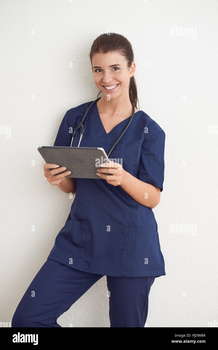 Giovane donna medico o infermiere in blu scrubs tenendo un tablet pc in piedi di fronte a un muro bianco Foto Stock
