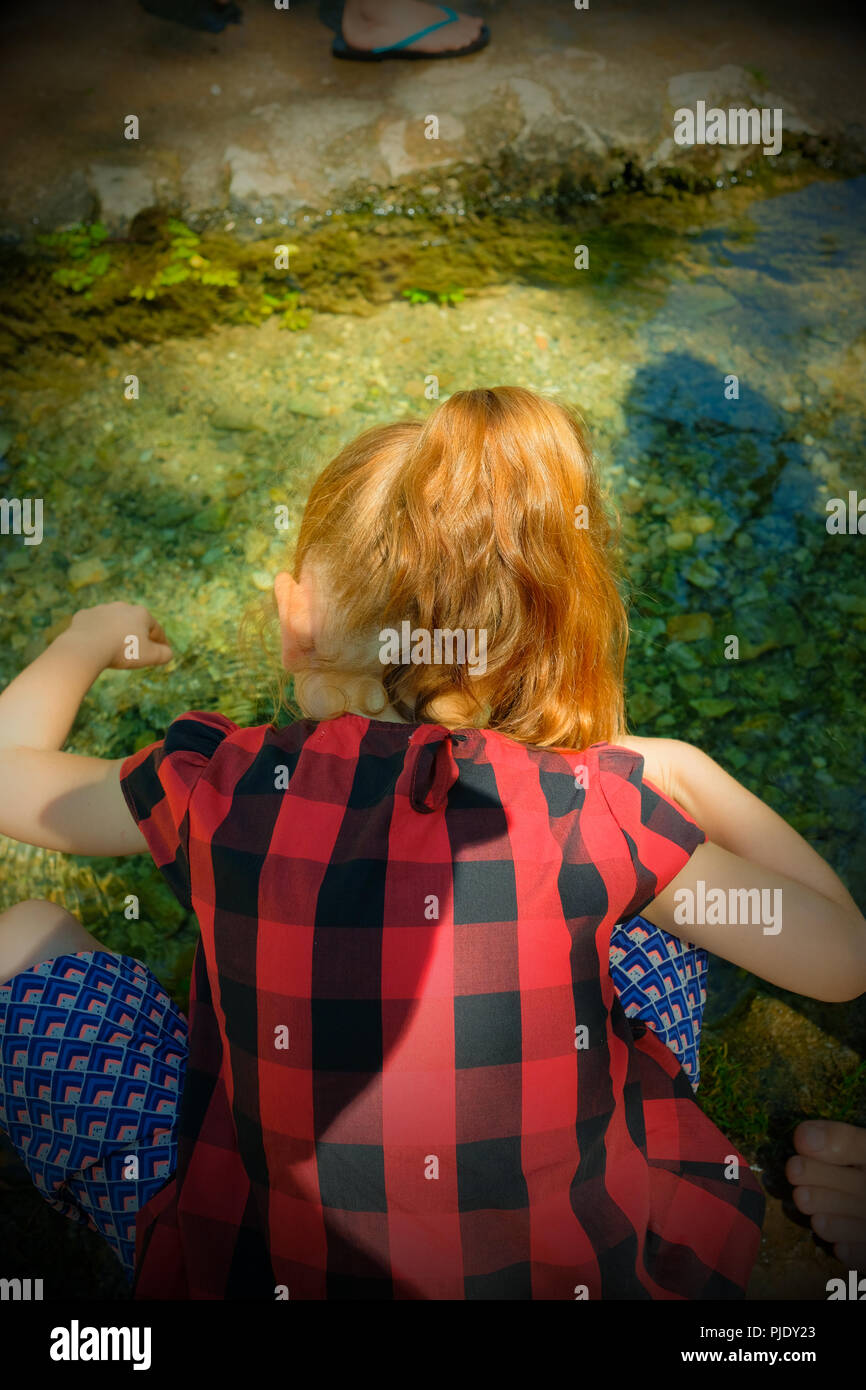 Vista posteriore di cinque anni ragazza francese con i capelli rossi accovacciato sul bordo di una banca di fiume guardando in giù nell'acqua con ombre o persona su acqua Foto Stock