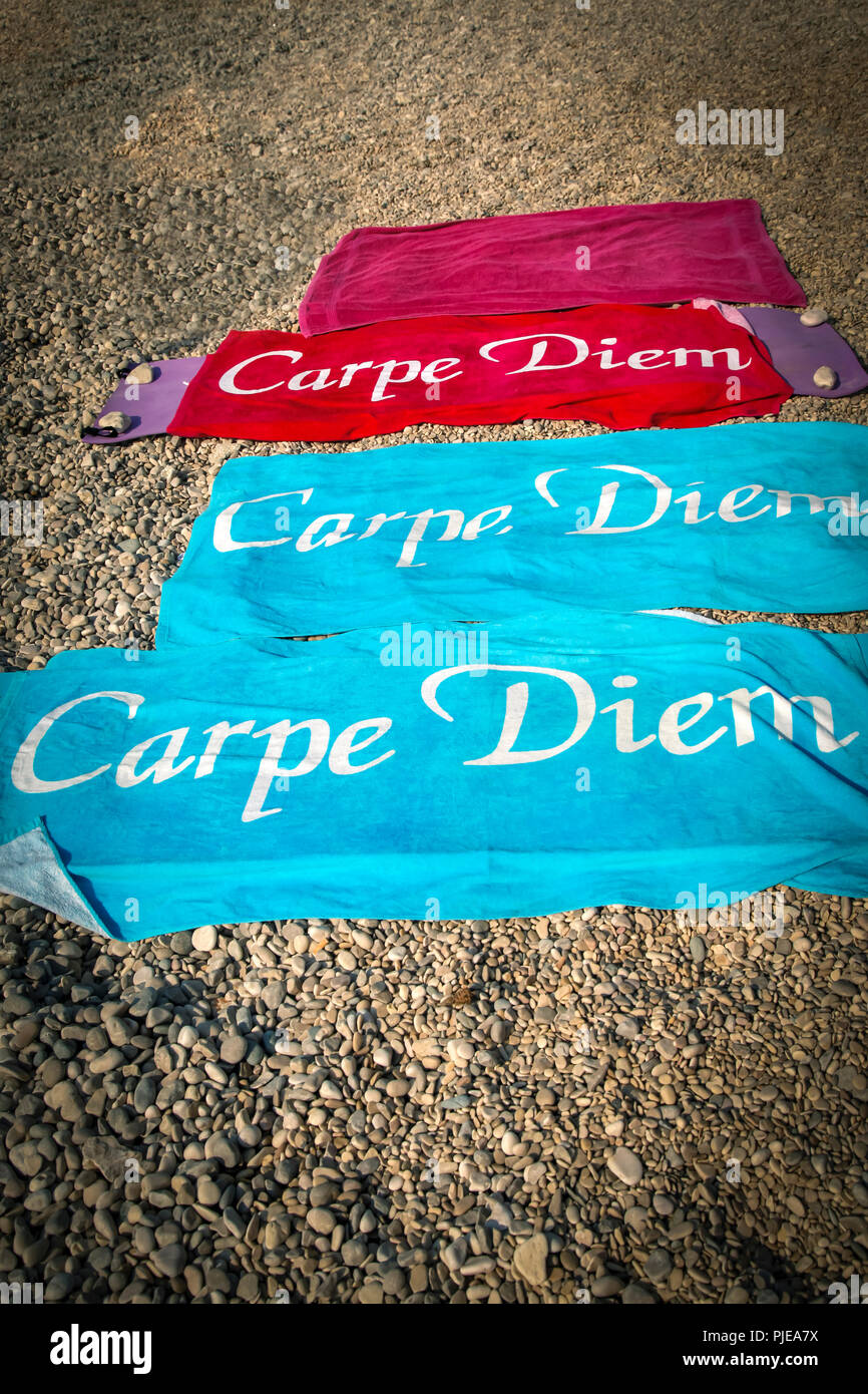 Tre coloratissimi asciugamani da spiaggia con la scritta "Carpe Diem", distesa su una spiaggia di ciottoli con rocce poste sui bordi per pesare giù dal vento Foto Stock