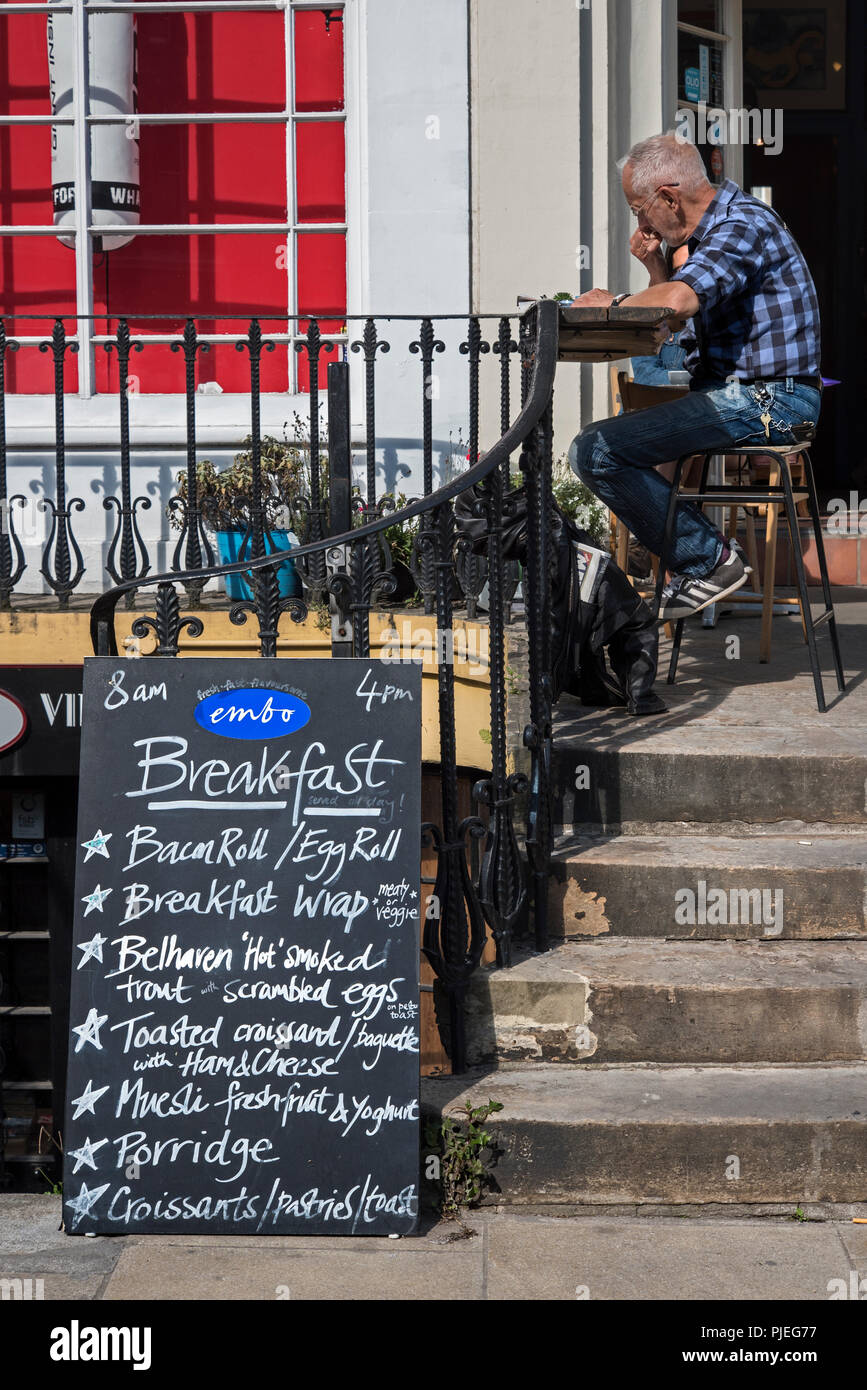 Un uomo maturo seduto sotto il sole al di fuori di un cafè sul Leith Walk di mangiare la prima colazione. Foto Stock