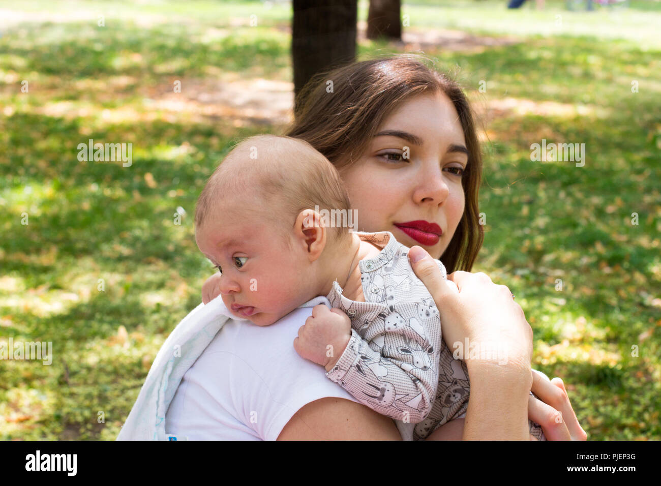 Giovane madre cura il suo grazioso baby sulla spalla al di fuori del parco durante la bella giornata di sole, infantili testa in appoggio sullo spallamento in posizione eruttazione Foto Stock