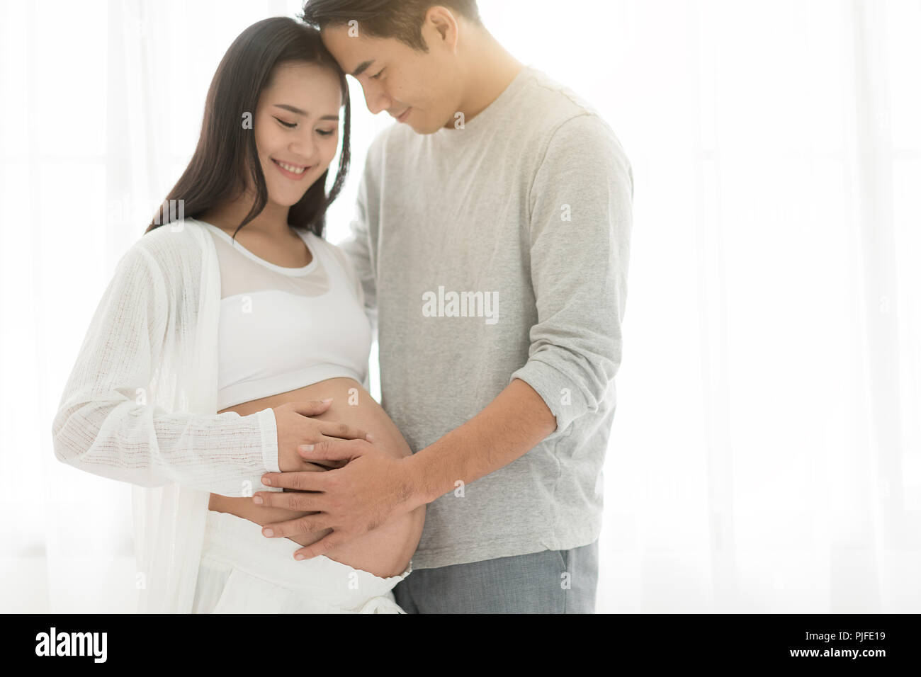 Bel uomo asiatico e la sua bella moglie incinta sono abbracciando e sorridenti mentre sta in piedi vicino alla finestra di casa. Foto Stock