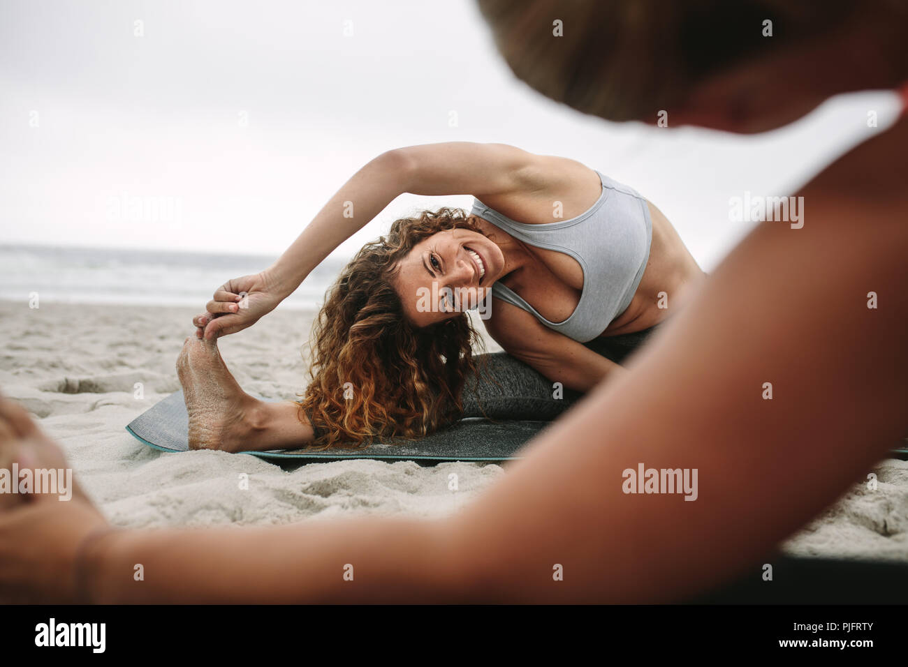 donne del fitness sedute su tappetini da yoga in spiaggia per allenarsi con lo stretching. Donne che praticano lo stretching posture di yoga sedute vicino al mare. Foto Stock