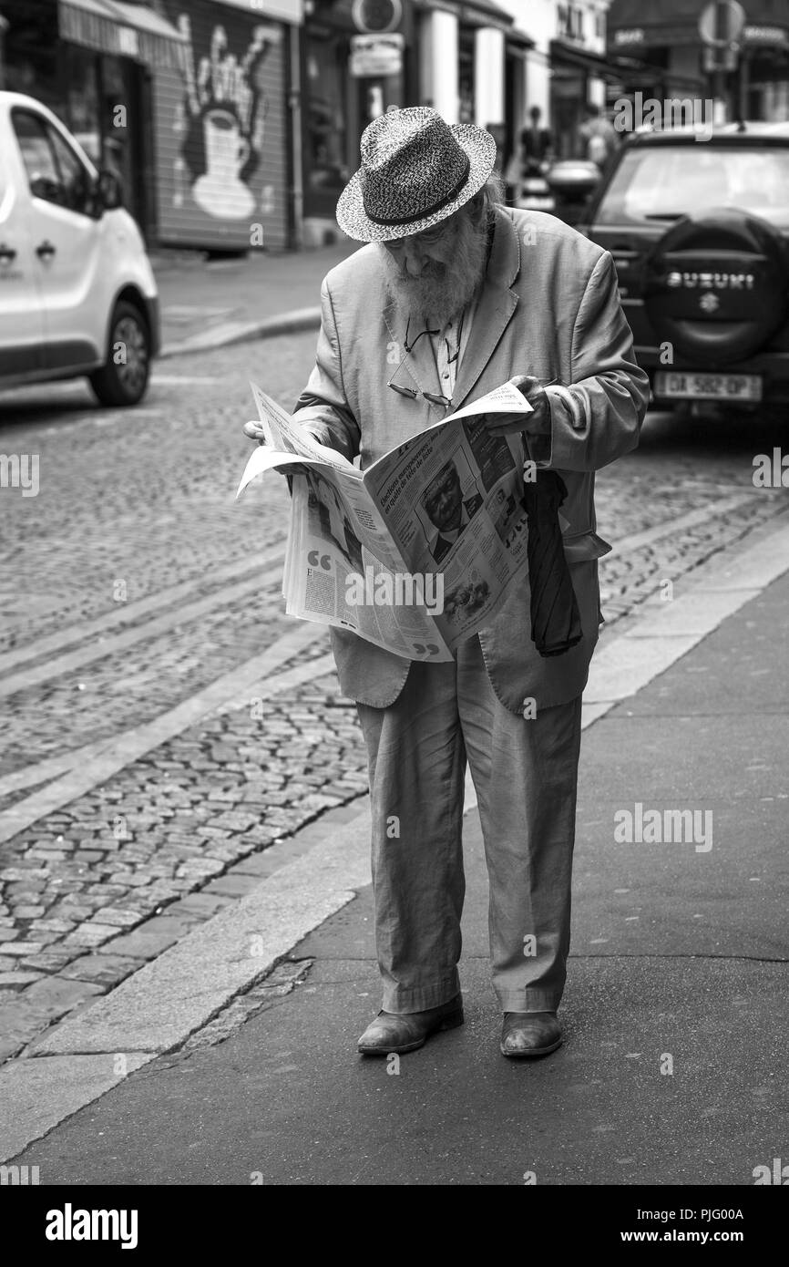 Un anziano gentiluomo barbuto che indossa un cappello e un abito, tenendo un ombrello e la lettura di un quotidiano su un marciapiede in Montmartre, Parigi Foto Stock