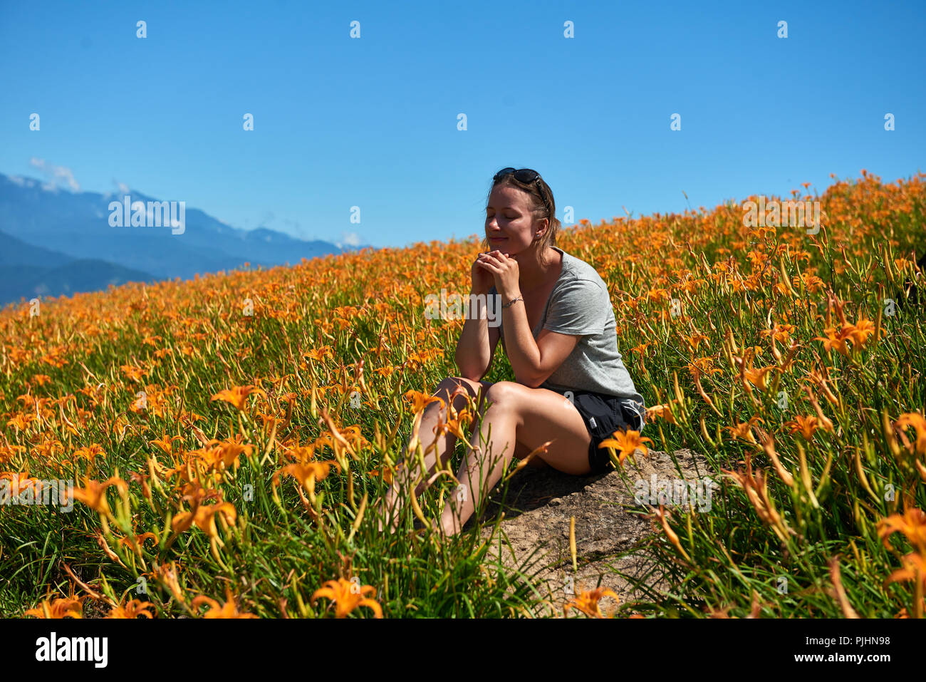 Giovane ragazza seduta nei campi di gigli arancione con le montagne sullo sfondo Foto Stock