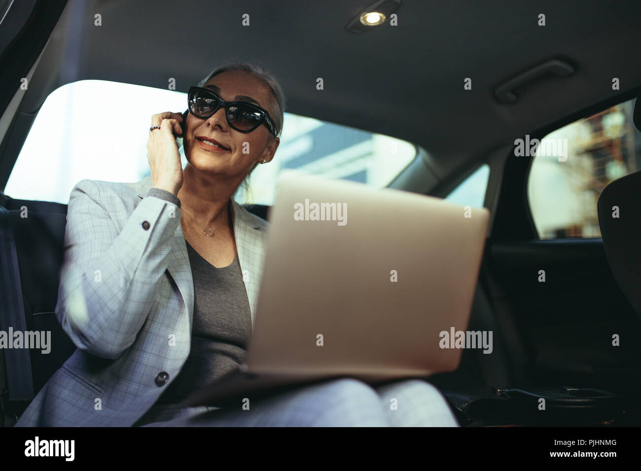 Imprenditore femmina in viaggio per ufficio in una lussuosa auto seduto sul sedile posteriore con il computer portatile e guardando fuori dalla finestra mentre parlano al telefono . Ma Foto Stock